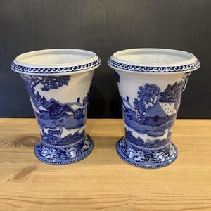 May include: A pair of white and blue ceramic vases. Each vase has a flared design with a detailed blue landscape scene. The rims and bases are decorated with a blue pattern. The vases are on a wooden surface.