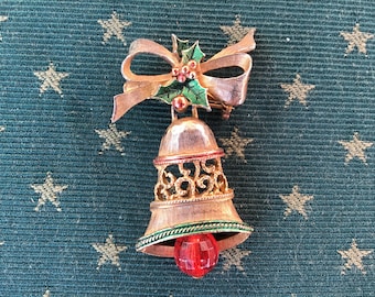 May include: A gold-tone Christmas bell brooch with a red jewel at the bottom. The bell is decorated with a green and gold bow and a green holly sprig with red berries.