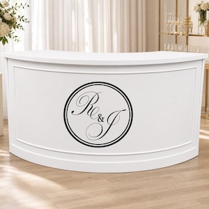 Op de afbeelding: Een witte, gebogen bar met een decoratief voorpaneel. Een zwart cirkelvormig embleem met de letters "Re & J" is gecentreerd op de voorkant. De bar staat tegen een achtergrond van bloemen en glaswerk.
