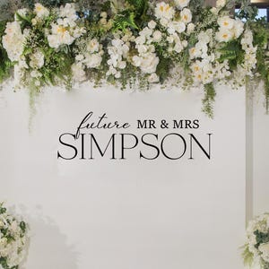 Può includere: Un muro bianco decorato con composizioni floreali e la scritta "future MR & MRS SIMPSON". L'immagine presenta una decalcomania in vinile ed è adatta per l'arredamento di matrimoni. Le composizioni floreali includono fiori bianchi e verde.