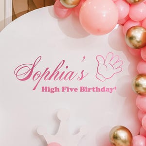 May include: A white circular backdrop with the text "Sophia's High Five Birthday" in pink. The backdrop is surrounded by pink and gold balloons. A white crown-shaped chair is in the foreground. A gray circle with the text "vinyl decal only" is in the bottom right.