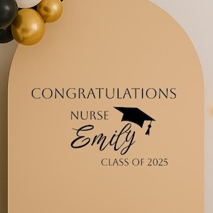 May include: A graduation party decoration with a black and gold balloon arch, a beige arch with the text "Congratulations Nurse Emily Class of 2025", a white cake with a black graduation cap, and a neon sign of a graduation cap.