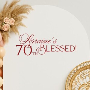 May include: A decorative party backdrop with a balloon arch in cream, peach, and bronze colors, adorned with pampas grass and roses. The text "Lorraine's 70th & BLESSED!" is displayed on a white arch. A rattan peacock chair is on the right.