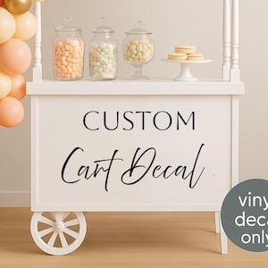 May include: A white cart with a decorative design, featuring the text "CUSTOM Cart Decal". The cart is decorated with balloons in shades of peach, cream, and gold. Glass jars filled with candies and cookies are displayed on the cart.