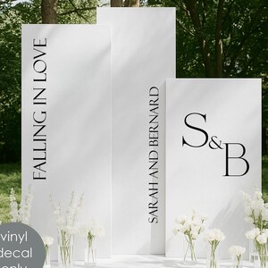 May include: A white wedding backdrop with black text reading "FALLING IN LOVE" and "SARAH AND BERNARD". The initials "S & B" are also displayed. White flowers in glass vases are arranged in front. A grey circle reads "vinyl decal only".
