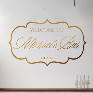 May include: Gold vinyl decal with a decorative frame and the text "Welcome to Michael's Bar Est. 2023". The decal is on a white wall above a bar counter with three wooden bar stools.