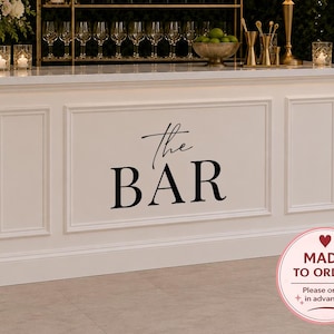 Puede incluir: Una barra blanca con el texto "the BAR" en negro. La barra tiene un estante de metal dorado con botellas de vino y copas. Una lámpara y un arreglo floral están en la barra. La imagen incluye el texto "VINYL DECAL ONLY" y "HIGH QUALITY VINYL DECAL".