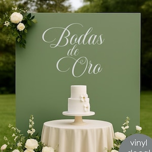 Op de afbeelding: Een witte bruidstaart met drie lagen en bloemdecoraties op een crèmekleurige tafel. De tafel staat voor een groene achtergrond met de woorden "Bodas de Oro" in witte letters. Een grijze cirkel zegt "vinyl decal only".