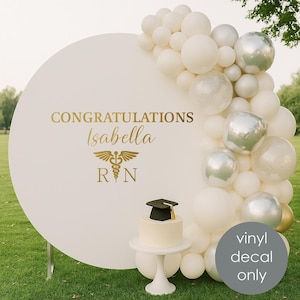 Peut inclure: Un panneau circulaire blanc avec le texte doré « Félicitations Isabella » et un symbole médical. Un petit gâteau avec un chapeau de graduation est posé sur un présentoir à gâteau blanc. Des ballons blancs et argentés décorent l'arrière-plan. Le texte « vinyle autocollant seulement » est sur un cercle gris.