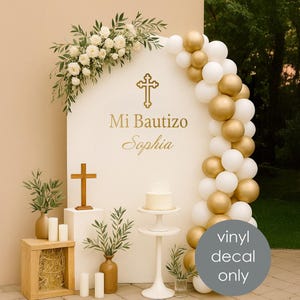 以下が含まれることがあります： 「Mi Bautizo Sophia」と金色の十字架が書かれた白いアーチ。アーチは白い花と緑で飾られています。金と白の風船のバルーンアーチが右側にあります。木の十字架とケーキが前にあります。