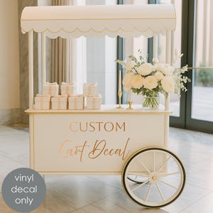 May include: A cream-colored custom cart with gold trim and a scalloped awning. The cart features the text "CUSTOM Cart Decal" in gold lettering. It is decorated with gift boxes, flowers, and candles. The cart has a large wheel.
