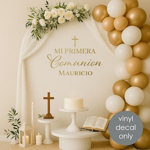 May include: A decorative backdrop for a First Communion celebration. The scene includes a white cake, a wooden cross, an open book, and balloons in gold and white. The text "MI PRIMERA Comunion MAURICIO" is displayed in gold.