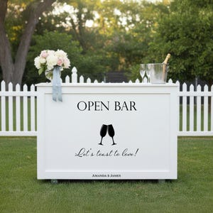 May include: White open bar with the text "OPEN BAR", two champagne glasses, and the phrase "Let's toast to love!". A bouquet of flowers and a champagne bottle in a bucket are on top. The bar is on wheels and sits on green grass.
