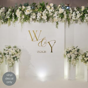 Puede incluir: Una pared blanca adornada con iniciales doradas y una fecha, rodeada de arreglos florales. Las iniciales doradas "W & Y" se muestran sobre la fecha "01.24.26". La imagen también incluye una pegatina "vinyl decal only".