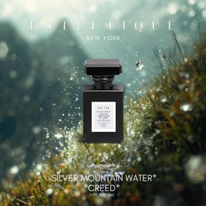 May include: A black perfume bottle with a rectangular shape and a square cap. The bottle has a white label with text that reads "Silver Mountain Water" and "Creed". The brand name "Esteetique" is at the top. The background is a blurred image of water and grass.