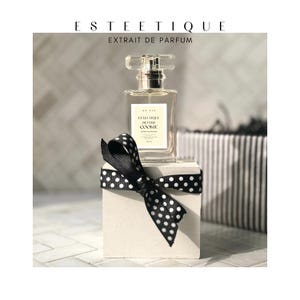 May include: A perfume bottle with a clear glass top and a rectangular body sits atop a small white cube adorned with a black ribbon featuring white polka dots. The bottle has a label that reads "ESTEETIQUE BUTTER COOKIE". The top of the image reads "ESTEETIQUE EXTRAIT DE PARFUM".