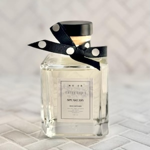 May include: A clear glass bottle with a black and white polka dot ribbon tied around the neck. The bottle contains a clear liquid and has a label that reads "No. 08 1st Hour SPEAKEASY REED DIFFUSER The art of fine fragrance and delicate blends of hand-selected essential oils. 100 ml."