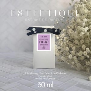 Puede incluir: Una caja de perfume blanca con un lazo de lunares blanco y negro. La caja tiene una etiqueta lila con el texto "LILAC" y "EXTRAIT DE PARFUM". Tambi&eacute;n se ve el texto "Introducing Lilac Extrait de Perfume". El frasco contiene 30 ml de perfume.