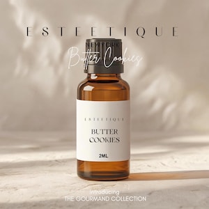 May include: A brown glass bottle with a black cap and a white label. The label reads "ESTETIQUE" and "BUTTER COOKIES" with "2ML" below. The text "Introducing THE GOURMAND COLLECTION" is at the bottom.