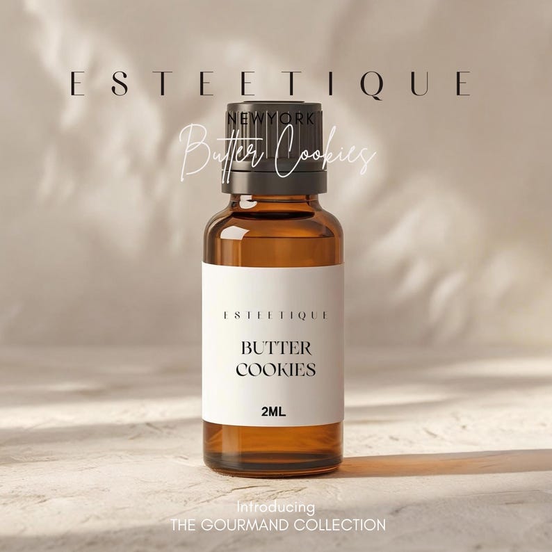 May include: A brown glass bottle with a black cap and a white label. The label reads "ESTETIQUE" and "BUTTER COOKIES" with "2ML" below. The text "Introducing THE GOURMAND COLLECTION" is at the bottom.