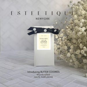 May include: A white rectangular box with a black and white polka dot ribbon. The box has a label that reads "ESTEE TIQUE BUTTER COOKIE". The text "Introducing BUTTER COOKIES #handcrafted HAUTE PARFUMERIE" is at the bottom.