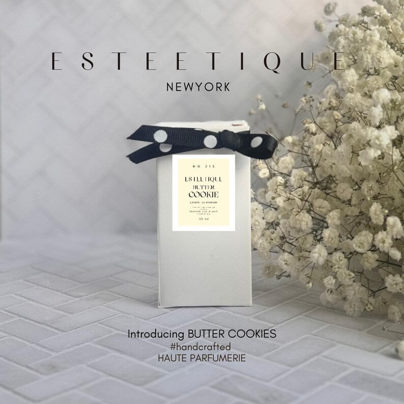 May include: A white rectangular box with a black and white polka dot ribbon. The box has a label that reads "ESTEE TIQUE BUTTER COOKIE". The text "Introducing BUTTER COOKIES #handcrafted HAUTE PARFUMERIE" is at the bottom.