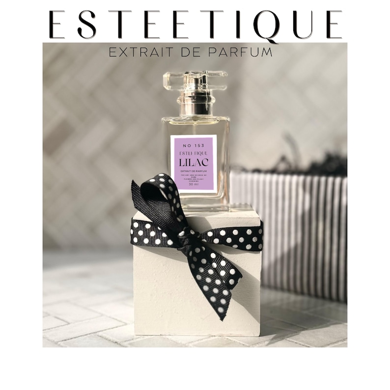 Puede incluir: Un frasco de perfume con un dise&ntilde;o de vidrio transparente y una etiqueta de color p&uacute;rpura claro que dice "LILAC". El frasco se exhibe sobre un bloque blanco adornado con un lazo de lunares en blanco y negro. El texto "ESTEETIQUE" est&aacute; en la parte superior.
