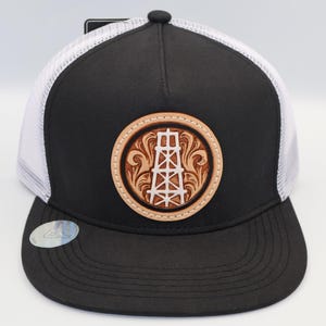 May include: A black and white trucker hat featuring a circular tan leather patch on the front. The patch displays an embossed oil derrick graphic surrounded by intricate scrollwork patterns. The hat has a solid black front panel and brim.