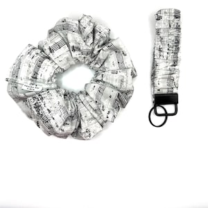 Op de afbeelding: Een wit en zwart gepatroneerd haar elastiekje en een bijpassende sleutelhanger met een zwarte metalen ring. Het patroon is muzieknoten en bladmuziek.
