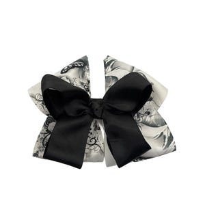 May include: A black and white hair bow. The bow features a large black bow on top of a patterned bow with a floral design. The bow is made of a grosgrain ribbon and is suitable for all ages.