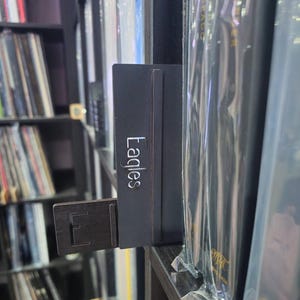 Puede incluir: Un soporte de álbum de vinilo negro con la palabra "Eagles" impresa en él. El soporte está unido a un estante con discos de vinilo.