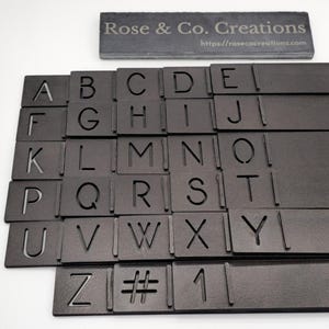 Puede incluir: Un conjunto de fichas de letras del alfabeto de metal negro con fondo negro. Las fichas están dispuestas en filas, cada fila contiene una letra diferente del alfabeto. Las fichas están hechas de metal y tienen un acabado liso y mate. Las letras están recortadas de las fichas, creando un efecto de plantilla.