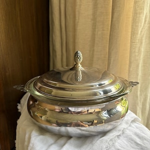 May include: A silver-plated serving dish with a lid and a decorative finial. The dish has a rounded base and two handles on either side. The lid is domed and reflects the surrounding environment. The dish is sitting on a white cloth.