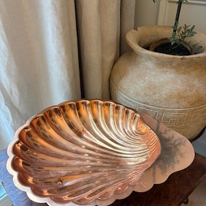 May include: A polished copper bowl shaped like a large seashell with deep, radiating fluted ridges. The metallic surface has a warm, reflective orange-brown tone and a scalloped edge, showcasing a distinct concave form for holding small items.