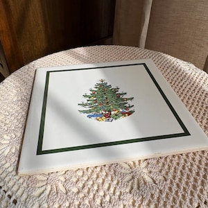 May include: A square, white decorative tile with a green border. The tile features a detailed illustration of a Christmas tree adorned with ornaments and surrounded by wrapped presents. The tile is placed on a cream-colored, crocheted surface.