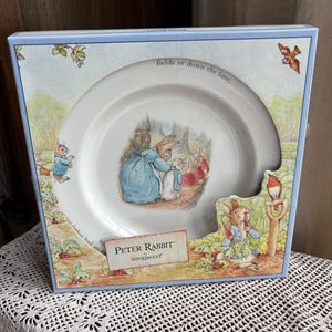 May include: A Peter Rabbit Wedgwood plate in a decorative box. The plate features a colorful illustration of Peter Rabbit and other characters in a forest scene. The box has a light blue border with additional illustrations of Peter Rabbit.