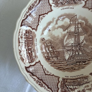 May include: Cream-colored ceramic bowl with a brown transferware design. The bowl features images of sailing ships, historical scenes, and decorative patterns. The rim has a scalloped edge and floral accents. Text includes "E Pluribus Unum", "Canton Port 1830", and "New York Harbor 1830".