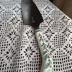 May include: A pie server with a dark, curved blade and a white handle. The handle is decorated with green holly leaves and red berries. The server is made of stainless steel and ceramic.