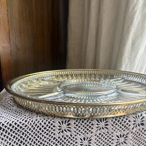 May include: A round serving tray featuring a clear glass center with a sunburst pattern, divided into sections. The tray is encased in a polished gold-tone metal rim with an intricate, openwork lattice design along the outer edge.