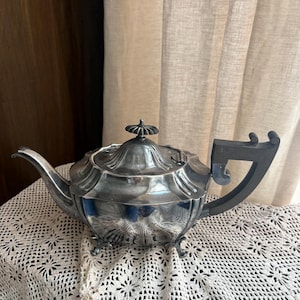 May include: A silver-colored teapot with a dark handle and a decorative lid. The teapot has a curved spout and sits on small feet. The teapot is placed on a white crocheted tablecloth.