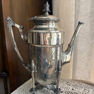 Könnte beinhalten: Eine silberfarbene antike Kaffeekanne mit einem dekorativen Blumendesign. Die Kanne hat einen Deckel, einen gebogenen Griff und einen Ausguss. Sie steht auf drei kleinen Beinen und steht auf einer weißen Spitzentischdecke.