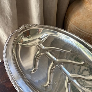 Puede incluir: Una bandeja de servicio ovalada de tono plateado con una superficie pulida y reflectante, y un patrón en relieve que recuerda a las venas de una hoja en el centro. El borde de la bandeja está decorado con un detalle texturizado de cuentas.