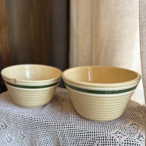 Puede incluir: Dos cuencos mezcladores de cerámica beige vintage con rayas verdes y blancas. Los cuencos están sobre un mantel de encaje blanco. Los cuencos tienen un diseño acanalado.
