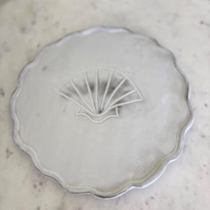 Könnte beinhalten: Ein weißer Keramikteller mit gewelltem Rand und einem Muscheldesign in der Mitte. Der Teller hat eine leicht strukturierte Oberfläche und eine dezente, cremefarbene Farbe. Das Muscheldesign ist erhaben.