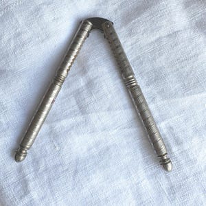 May include: Silver-coloured nutcracker with a textured, ridged design. The nutcracker is open, showing its hinged mechanism. The handles have decorative bands and rounded ends. It rests on white, textured fabric.