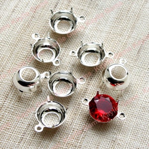 May include: Eight silver-toned metal settings for cabochon stones, with one setting featuring a red cabochon stone.
