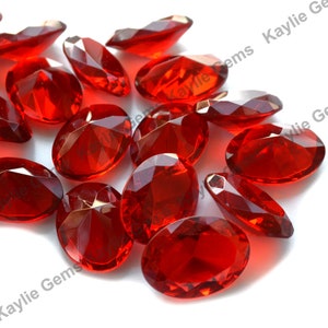 May include: A group of 16 red oval-shaped faceted glass gems. The gems are scattered on a white surface.