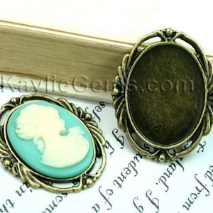 Könnte beinhalten: Zwei antike, goldfarbene Metall-Cameo-Fassungen. Eine Fassung ist leer, die andere hat einen hellblauen und weißen Cameo-Einsatz. Der Cameo-Einsatz zeigt das Profil einer Frau.