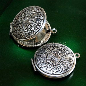 May include: Two silver locket pendants with floral designs. The lockets are open and reveal a mirror inside. The lockets are on a green background.