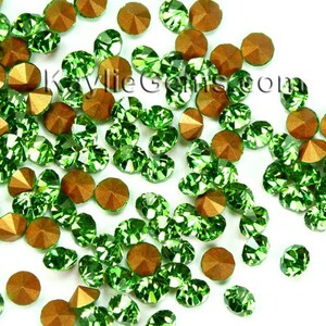 May include: Assortment of green and gold rhinestones. The rhinestones are scattered on a white background.